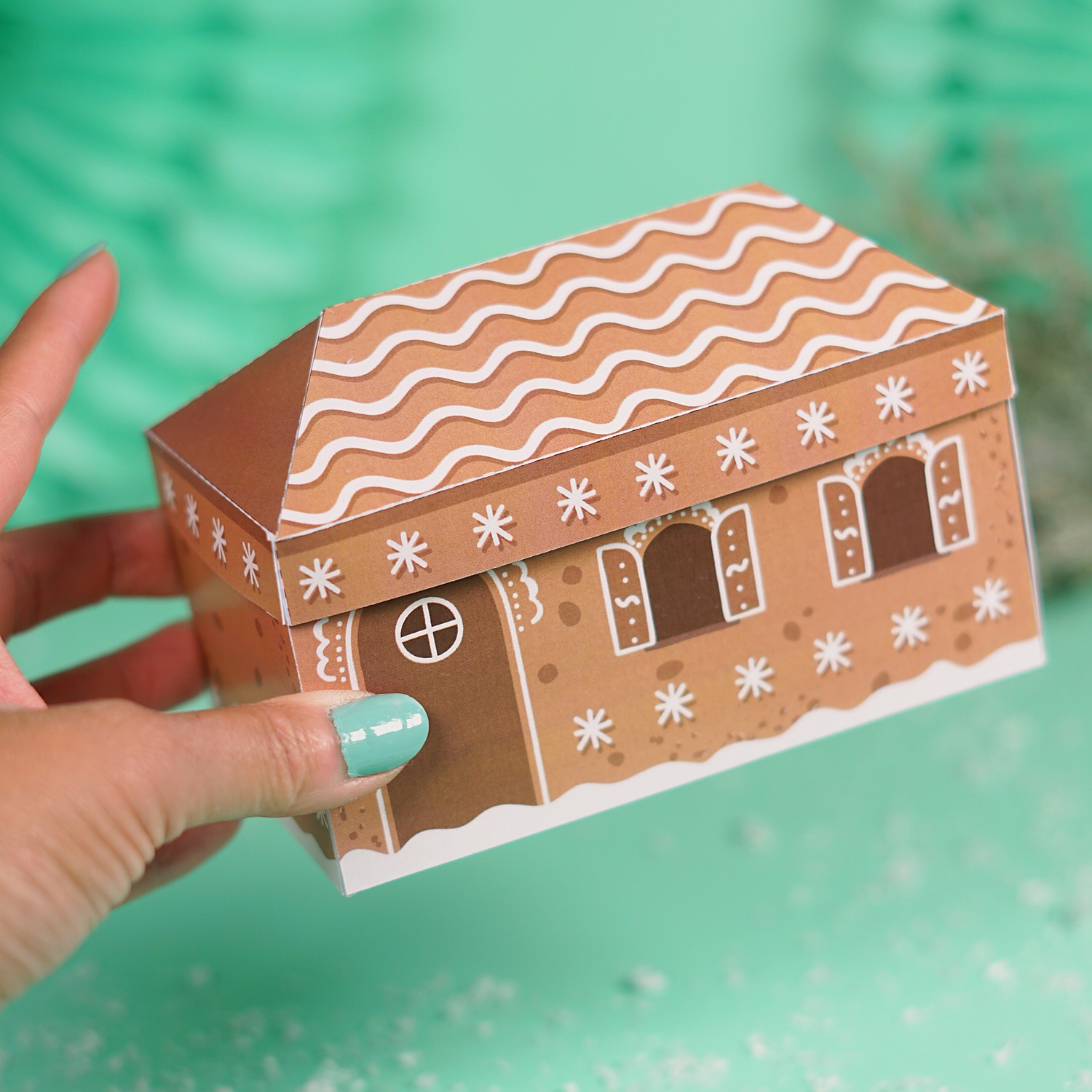 Zwei Hände halten das fertige Lebkuchenhaus Geldgeschenk mit Zuckergussdach vor mintfarbenem Hintergrund
